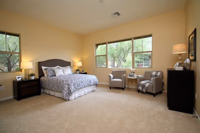 Spacious bedroom with a bed, two armchairs by a large window, nightstands and soft neutral decor.