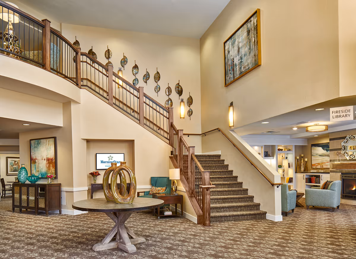 Interior view of a senior living facility lobby featuring a carpeted staircase with wooden railings, a round table with decorative sculptures in the center, comfortable seating areas with armchairs, wall art, and a sign indicating the Fireside Library near a fireplace.