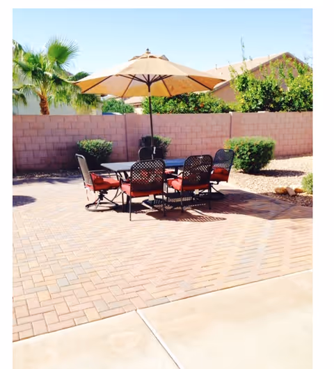Outdoor patio area with a table and six chairs with red cushions under a large beige umbrella. The patio is paved with bricks and surrounded by a low brick wall, with green bushes and palm trees visible in the background under a clear blue sky.