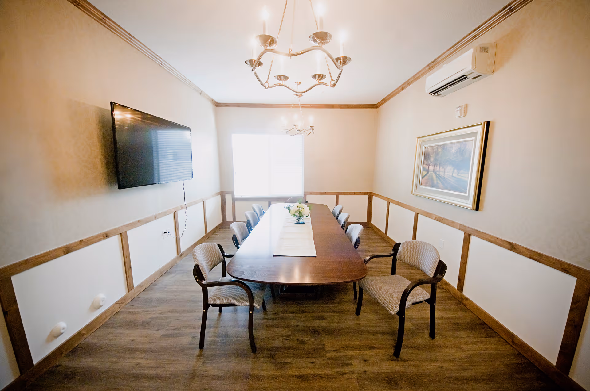 A dining room with a long wooden table surrounded by eight chairs. The room has wooden flooring, beige walls with wooden trim, a wall-mounted flat screen TV, a framed landscape painting, two chandeliers, and a window letting in natural light. An air conditioning unit is mounted on the wall.
