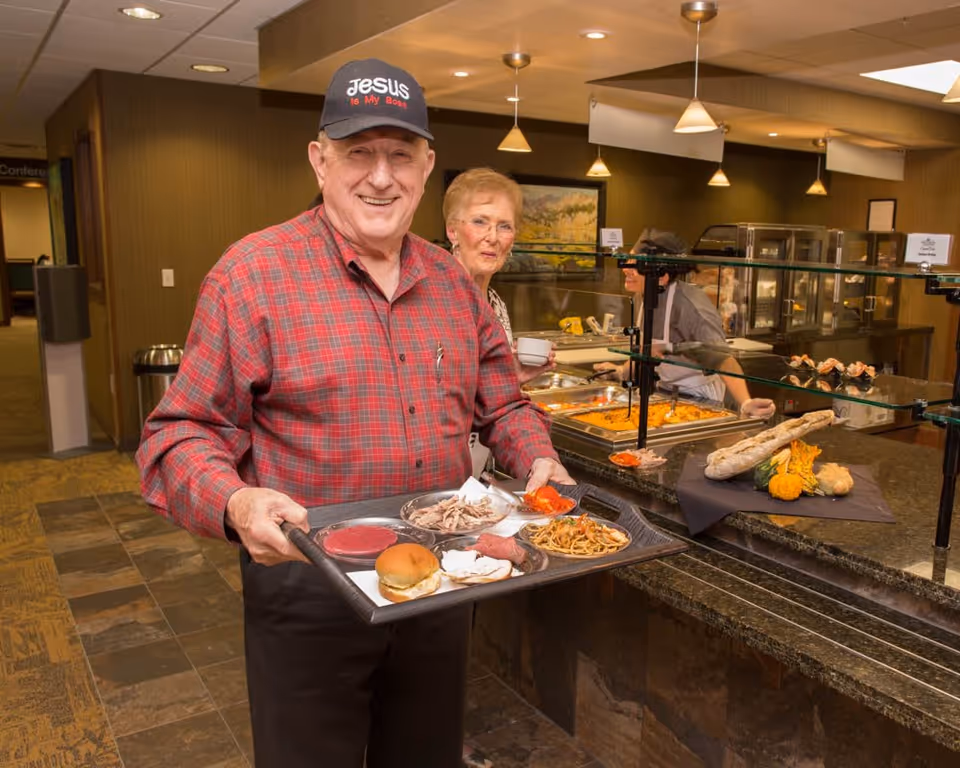 An elderly man wearing a red plaid shirt and a black cap that says 'Jesus is My Boss' is holding a tray with various foods including a sandwich, noodles, and sliced meats in a cafeteria-style dining area. Behind him, an elderly woman is holding a cup, and a cafeteria worker is serving food behind a glass counter with trays of hot dishes.