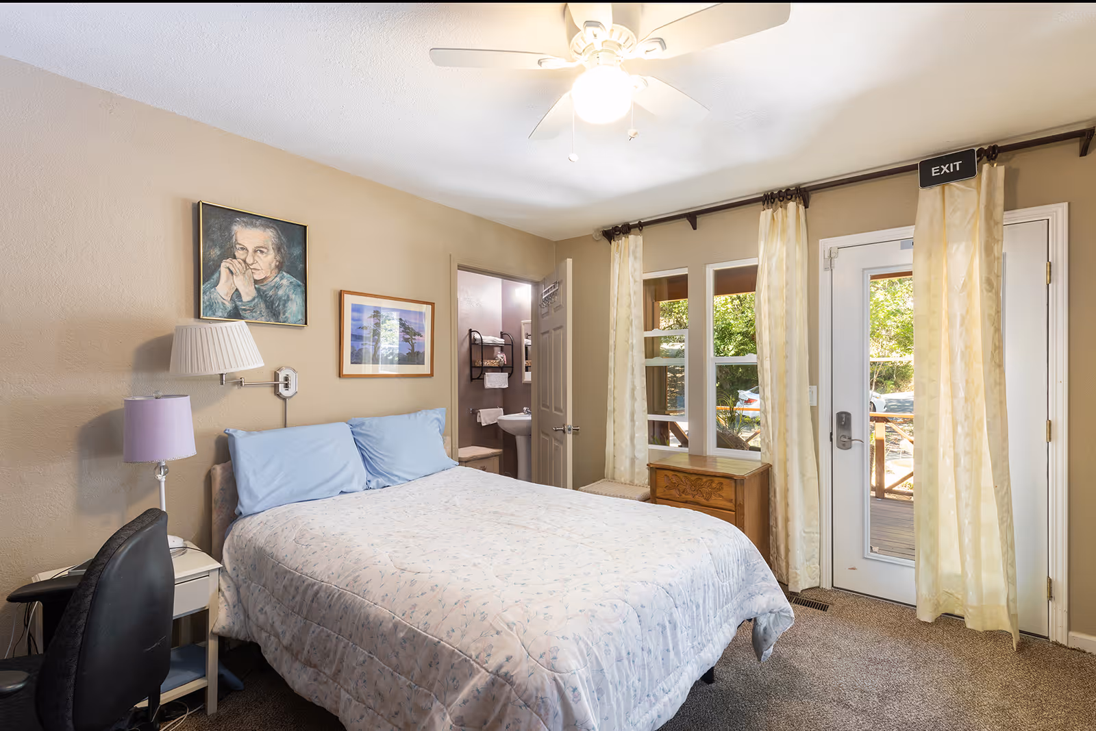 Bedroom with a bed, bedside lamps and desk chair, an open doorway to a bathroom, and a glass door with curtains leading outside.