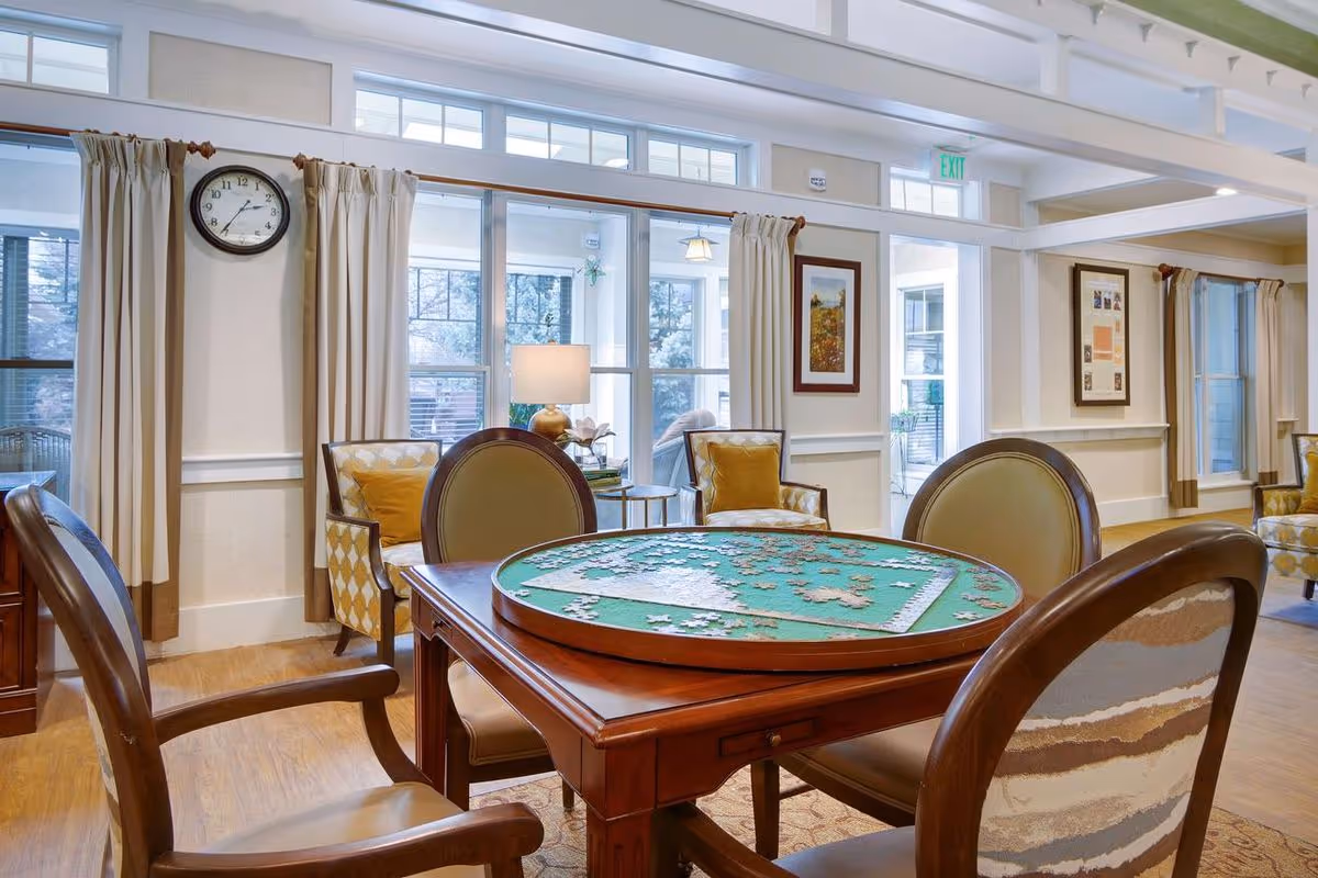 Well-lit senior living common room with a round table holding a partially completed jigsaw puzzle and chairs around it.
