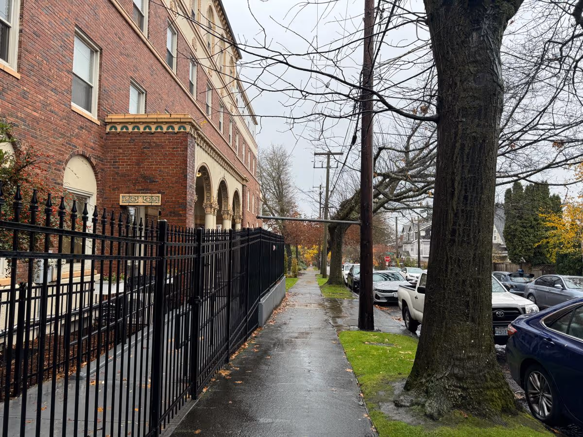 Brick multi-story building with arched entrance and a black iron fence along a wet sidewalk lined with trees and parked cars.