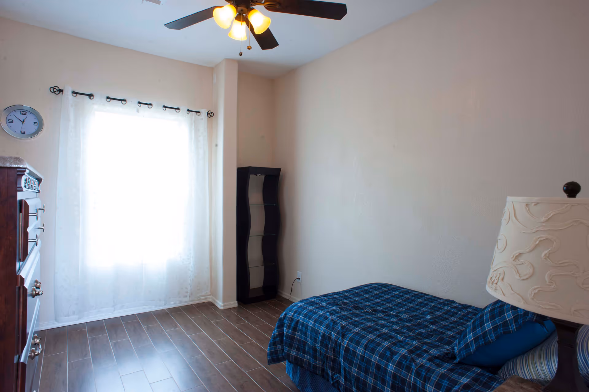 A simple bedroom with a single bed covered in blue plaid bedding, a wooden dresser, a small black curved shelf, a window with sheer white curtains, a ceiling fan with lights, and a decorative table lamp.