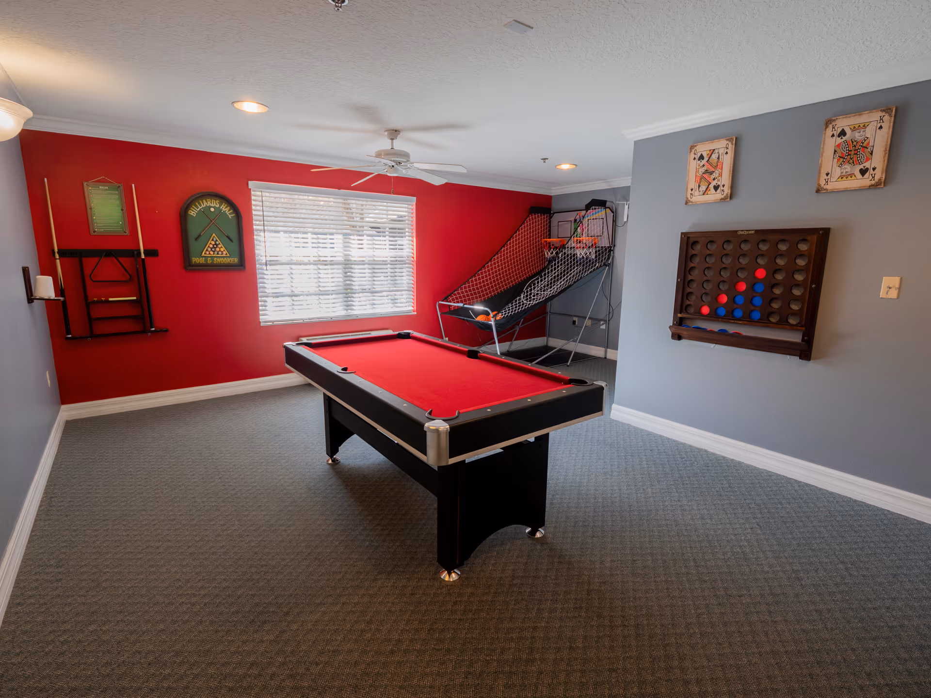 A game room with a red pool table in the center, a basketball arcade game in the corner, and a wall-mounted Connect Four game. The walls are painted red and gray, with a window letting in natural light. There are also two framed playing card pictures and a rack holding pool cues and accessories.