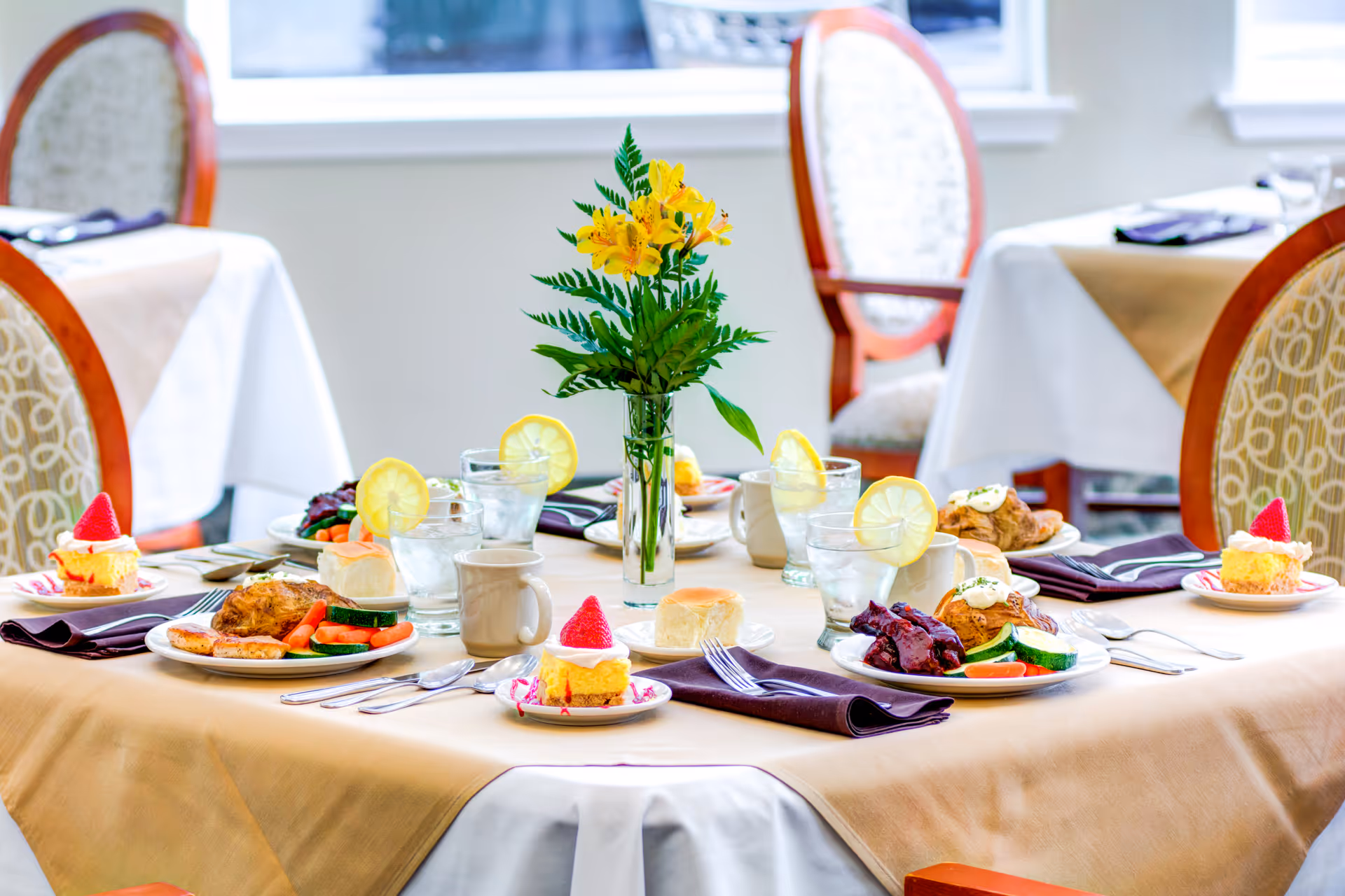 A dining table set for a meal with plates of food including chicken, vegetables, rolls, and dessert topped with strawberries. There are glasses of water with lemon slices, coffee mugs, and purple napkins. A vase with yellow flowers is placed in the center of the table. The background shows upholstered chairs and windows letting in natural light.