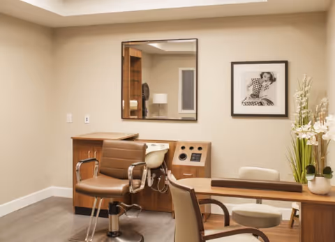 Interior of a salon or grooming room with a brown salon chair, a wooden counter with a built-in hair washing sink, a mirror on the wall, a framed black and white photo, and a table with chairs and a vase of flowers.