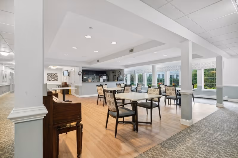 A bright and spacious dining area in a senior living facility with several tables and chairs arranged on a wooden floor. Large windows on the right side allow natural light to fill the room, and a counter area is visible in the background. The space is clean and well-lit with recessed ceiling lights, and there is a piano near a white column in the foreground.