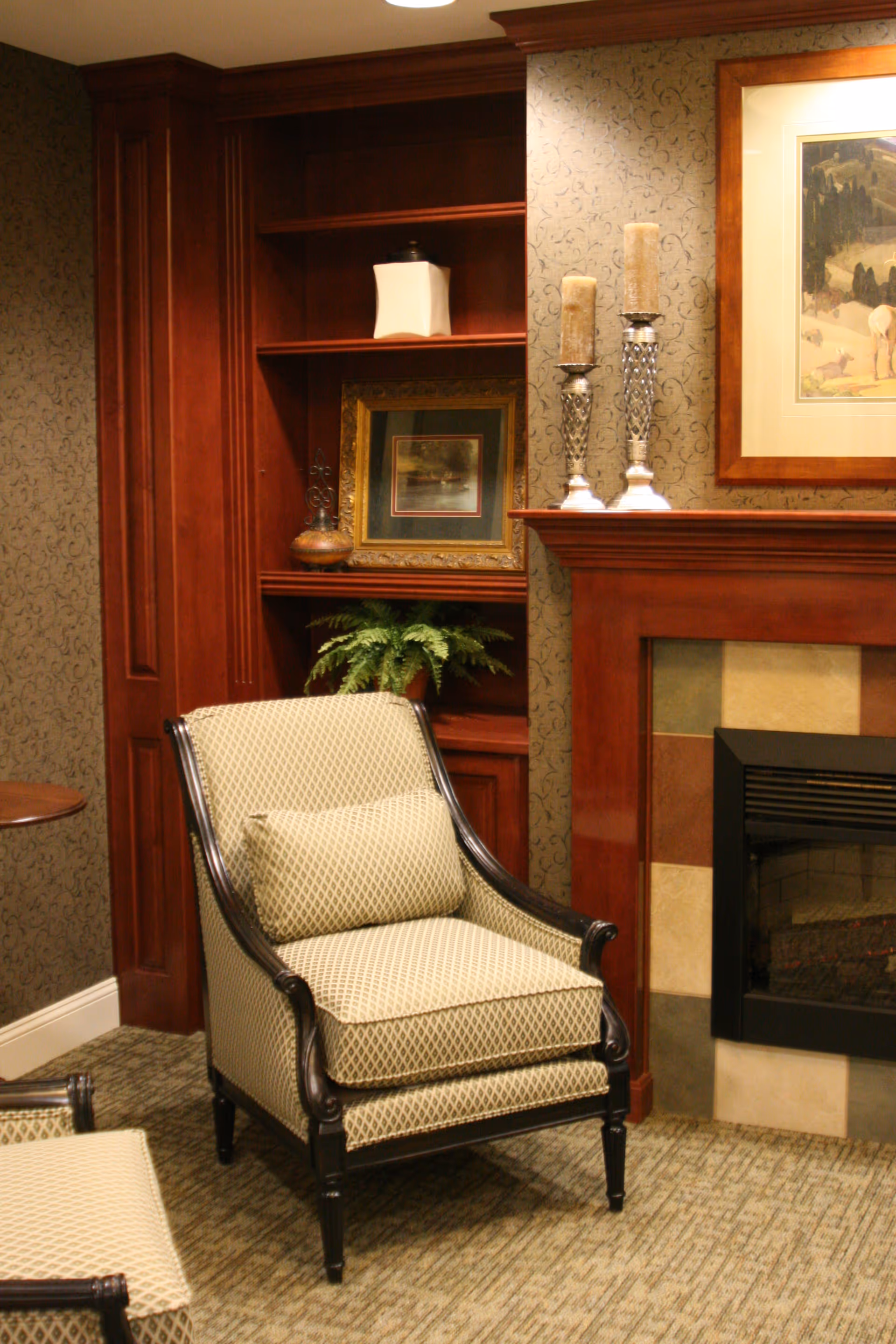 A cozy living room corner featuring a patterned armchair with a matching cushion, a wooden built-in bookshelf with decorative items including a framed picture and a plant, and a fireplace with a tiled surround and two tall candles on the mantel. A framed painting hangs above the fireplace.