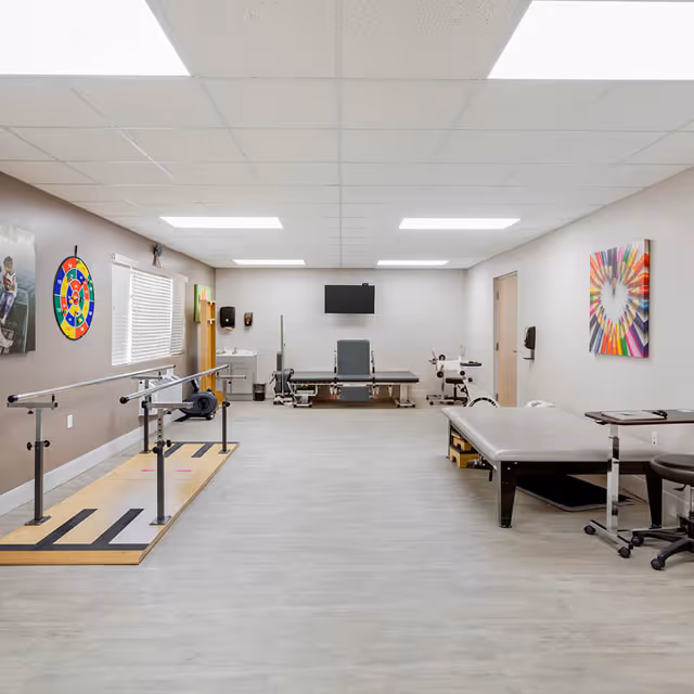A bright physical therapy room with parallel bars, treatment tables, exercise equipment, and wall art.