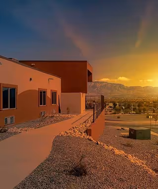 Exterior view of a building at sunset with a paved walkway, rocky landscaping, and mountains in the background under a colorful sky.