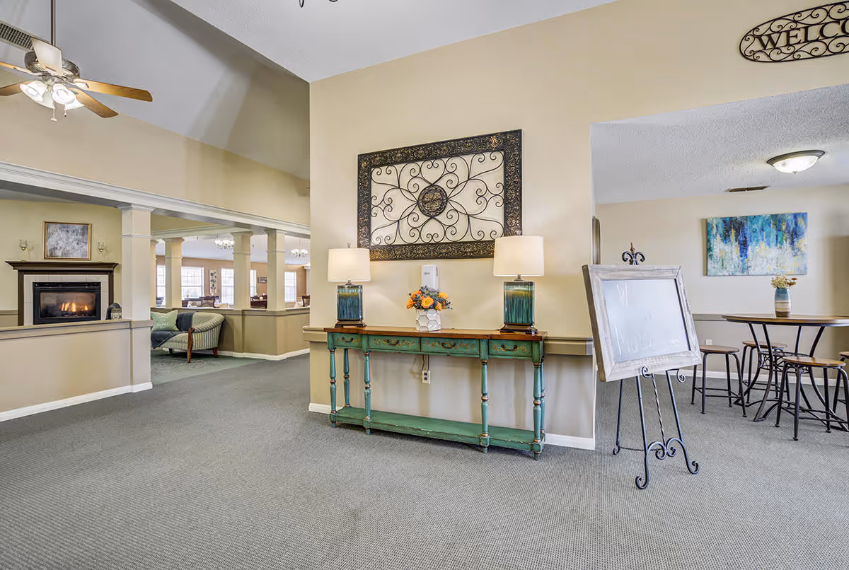 Interior view of a senior living facility lobby area with a green console table holding two lamps and a flower arrangement. Above the table is a decorative wrought iron wall art. To the left, there is a seating area with a fireplace and armchairs. To the right, there is a small dining area with a round table and stools, and a colorful abstract painting on the wall.