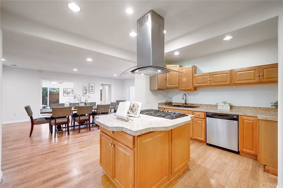 A modern kitchen with wooden cabinets, a central island with a gas stove and a stainless steel range hood. The kitchen has a dishwasher, a sink, and marble countertops. In the background, there is a dining area with a wooden table and eight upholstered chairs, with a sliding glass door leading to an outdoor space.