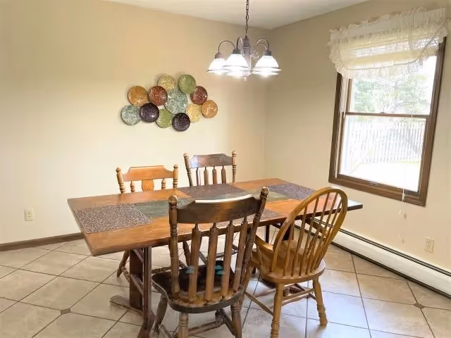 A dining room with a wooden table and four mismatched wooden chairs. The table has four placemats and a decorative wall hanging made of colorful plates. There is a window with sheer curtains letting in natural light, and a ceiling light fixture with five bulbs above the table. The floor is tiled and the walls are painted beige.