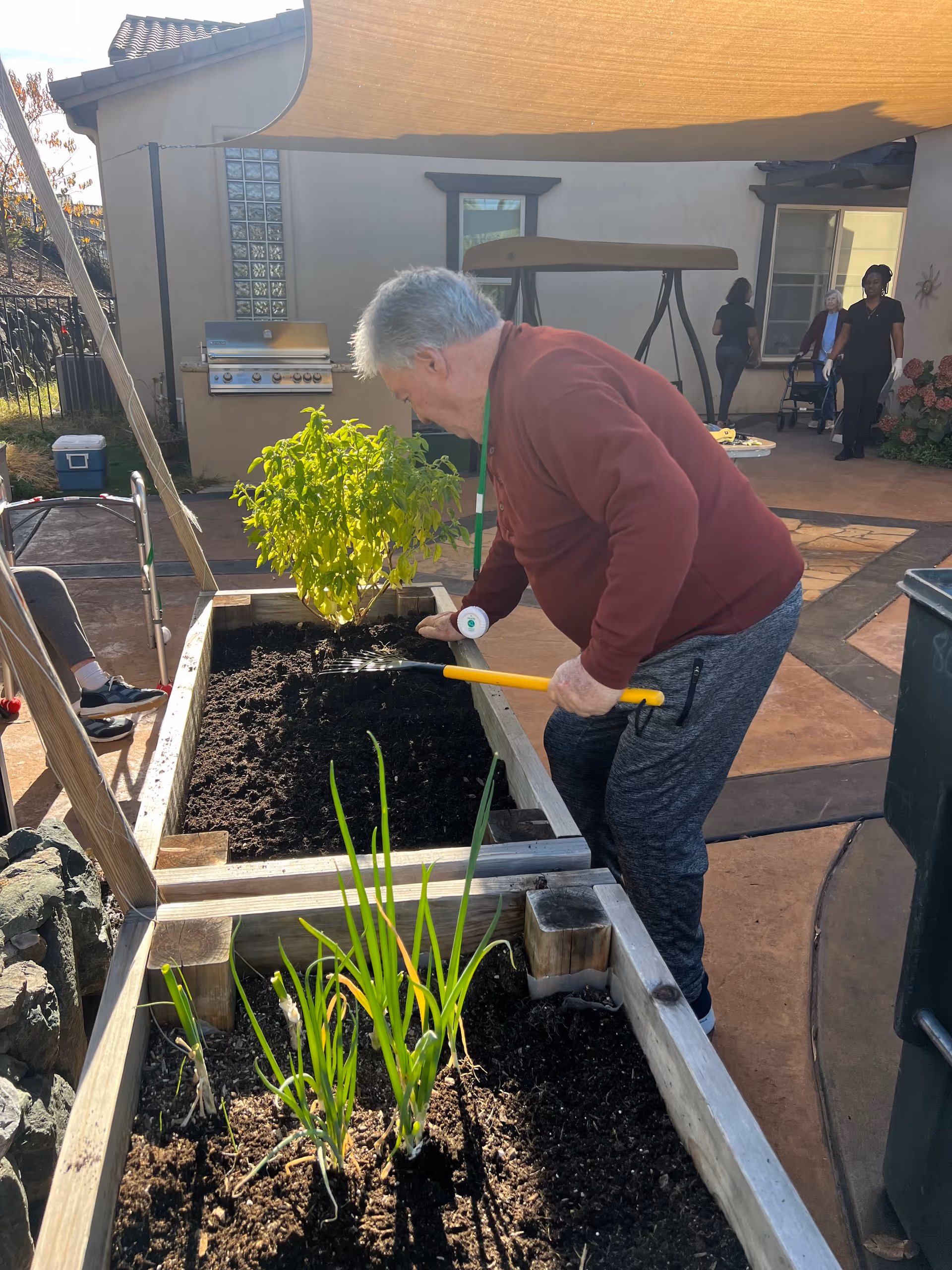 A person tends a raised garden bed in an outdoor courtyard, using a hand tool while others stand near the building in the background.