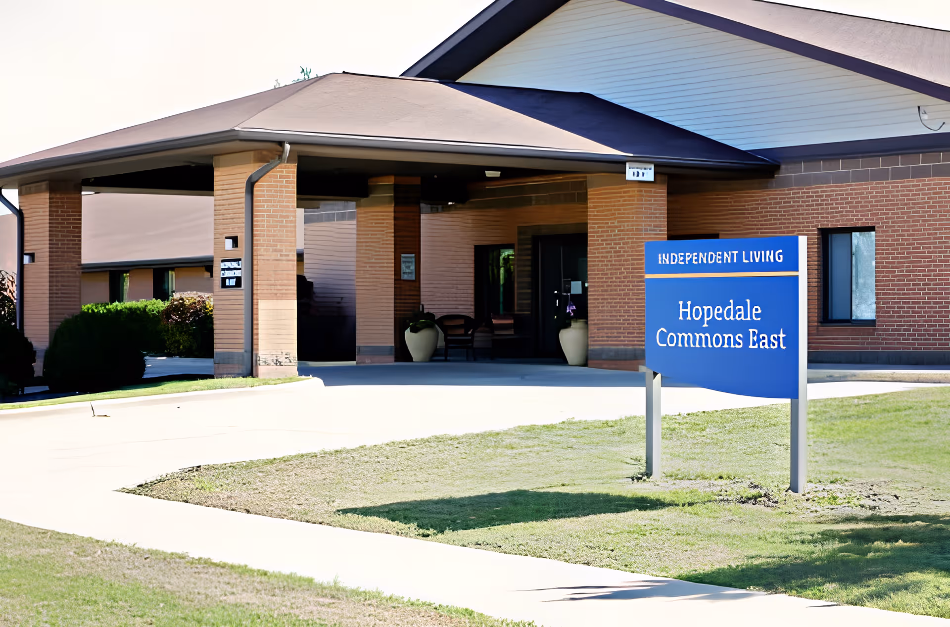 Front entrance of a brick senior living building with a covered drop-off, driveway, and a blue sign reading 'Hopedale Commons East'.