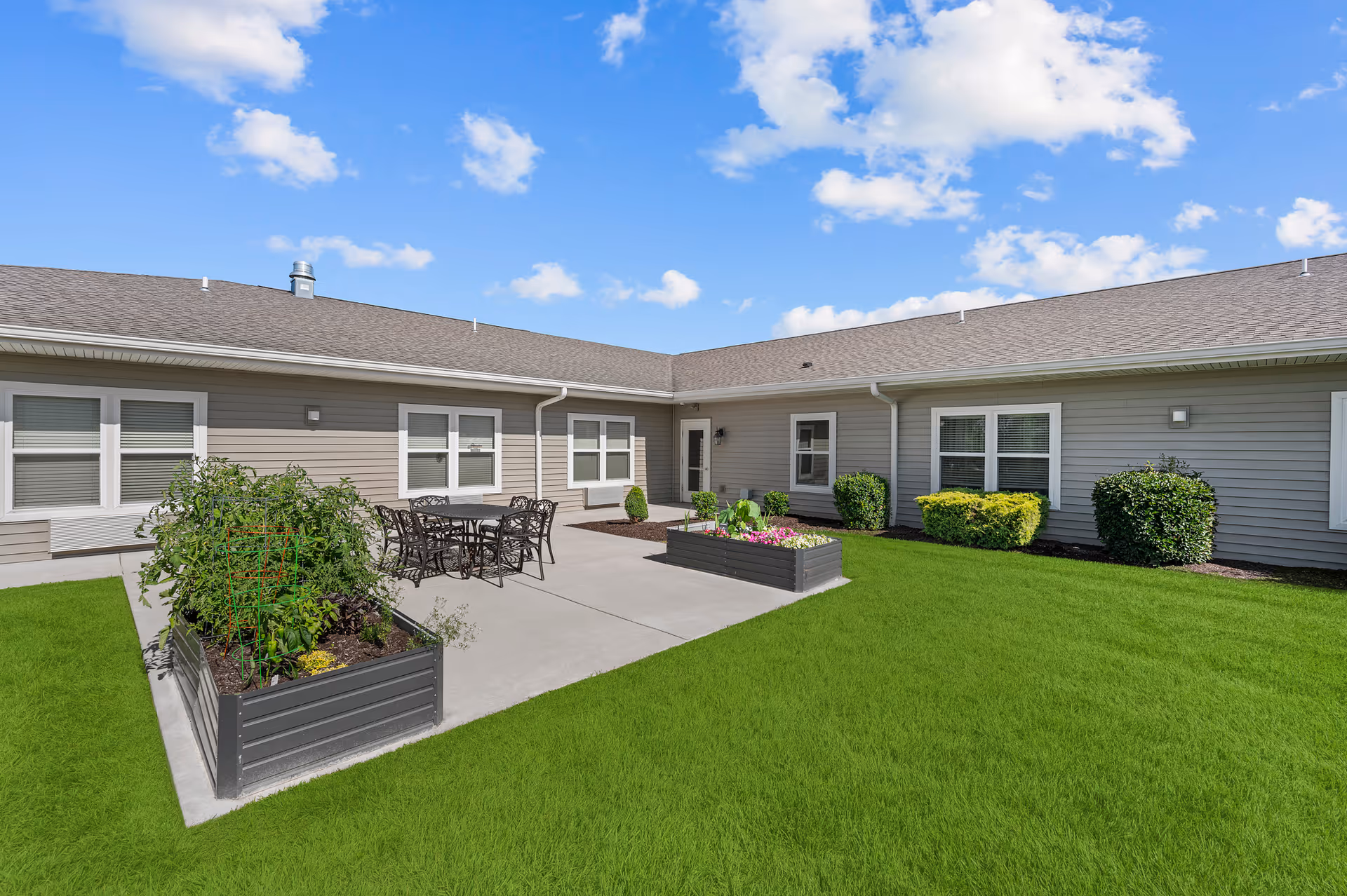 Outdoor courtyard area at Harton Senior Living with green grass, two raised garden beds containing plants, a concrete patio with a black metal table and chairs, and the exterior of a single-story building with multiple windows under a blue sky with scattered clouds.