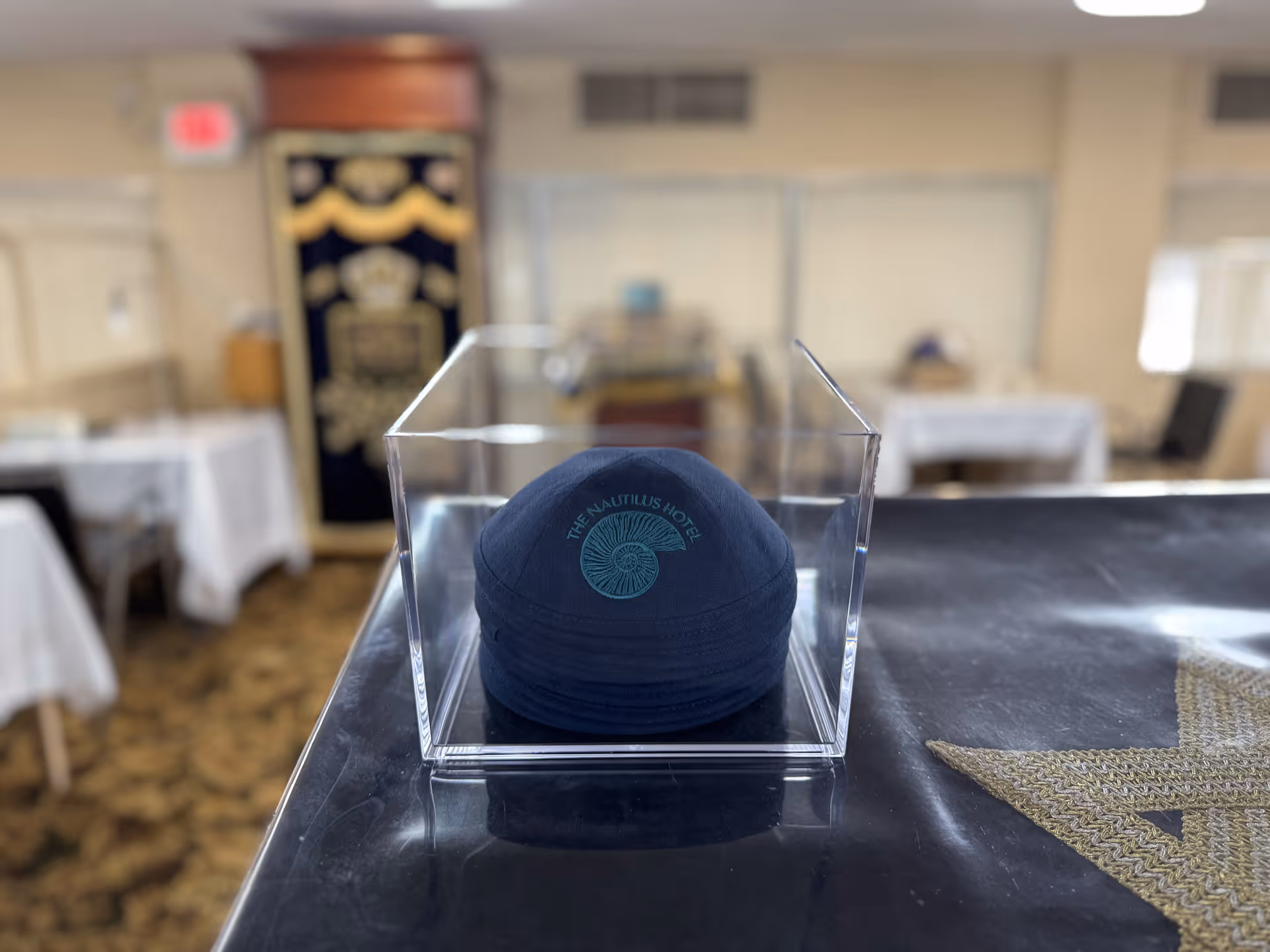A stack of dark blue kippahs with the logo and text 'The Nautilus Hotel' displayed inside a clear acrylic box on a dark surface, with a blurred background showing tables covered with white tablecloths and a decorative curtain with gold and black patterns.