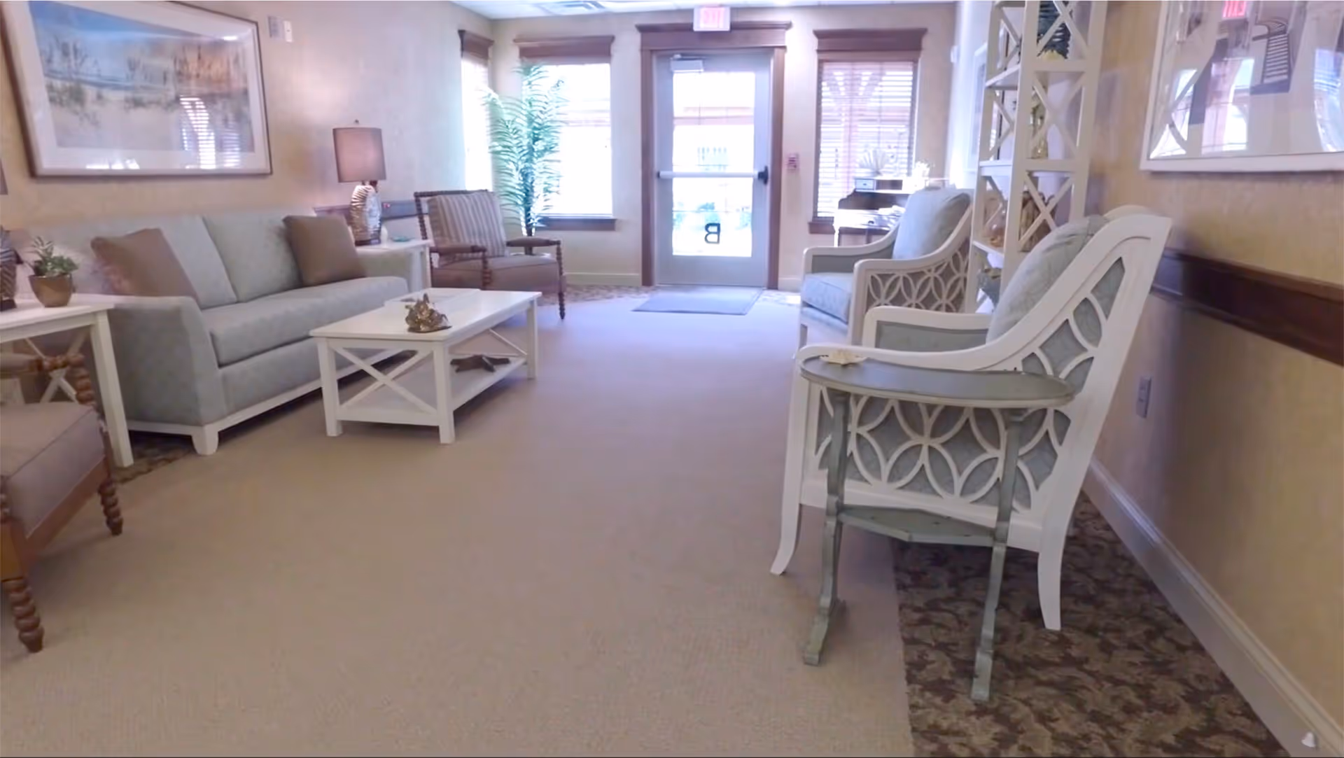 A cozy living room area with light blue upholstered sofa and chairs, a white coffee table, side tables with lamps, framed artwork on the walls, and a glass door with windows letting in natural light.
