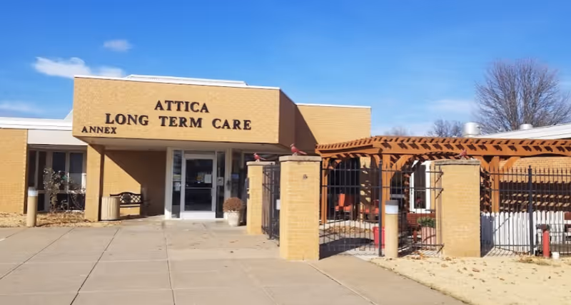 Exterior view of Attica Long Term Care Facility Annex building with a brick facade, entrance doors, a bench, and a fenced outdoor seating area with a wooden pergola under a clear blue sky.