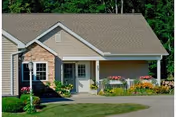 Exterior view of a single-story residential building with beige siding and a stone accent wall near the entrance. The building has a gable roof and a small porch area with a white door and windows. There are flower beds with colorful flowers and greenery along the front, and a paved driveway leading up to the entrance. Trees and greenery are visible in the background.