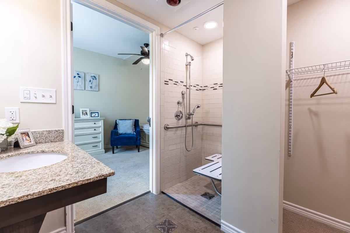 View of a bathroom area with a granite countertop and sink on the left, an open walk-in shower with a fold-down bench and grab bars in the center, and a closet space with a hanging rod and a single wooden hanger on the right. Through the open door, a bedroom is visible with a blue armchair, a white dresser, and wall art above the dresser.
