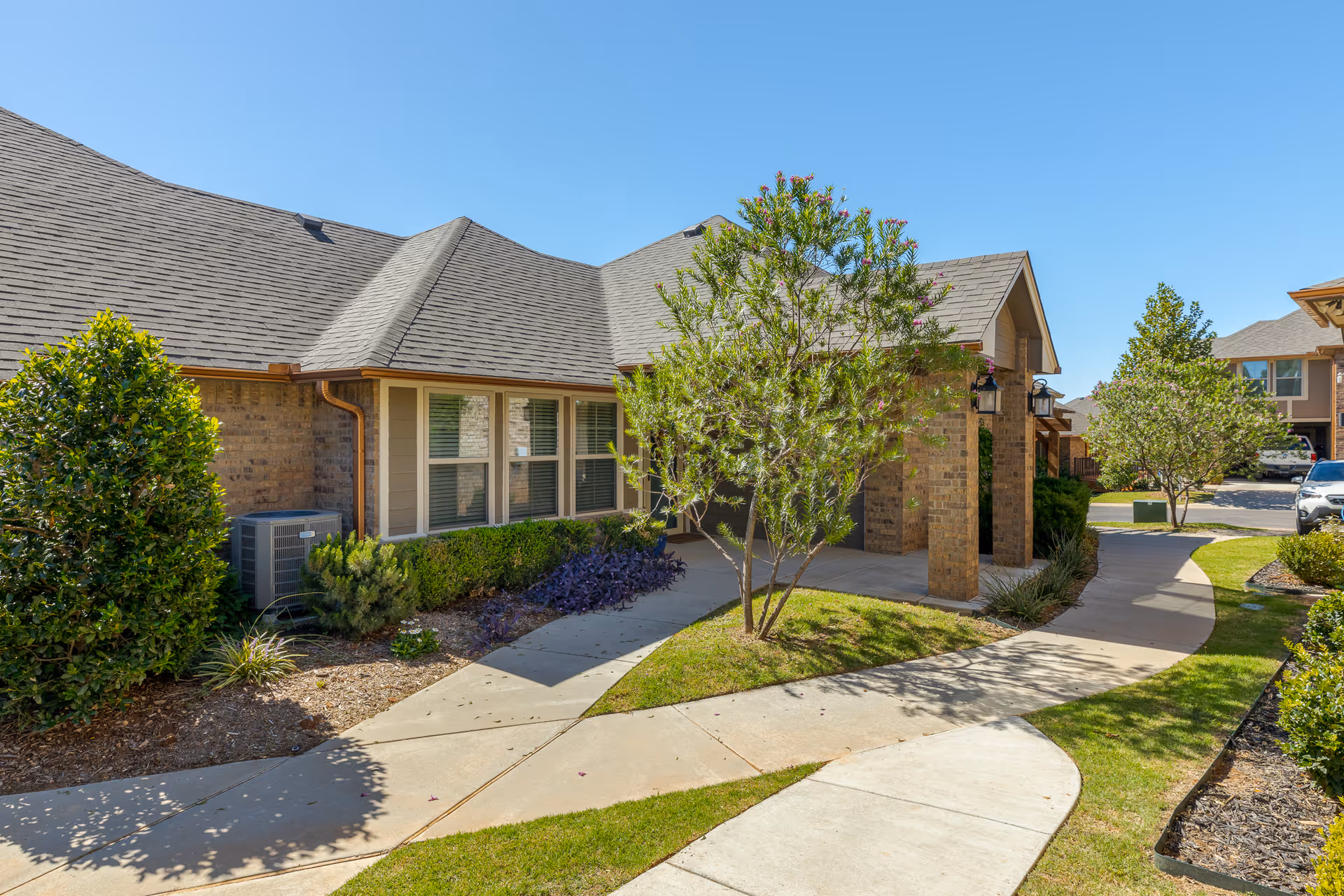 Exterior view of a single-story brick building with a sloped roof, surrounded by a well-maintained garden with shrubs and small trees, and a curved concrete walkway leading to the entrance under a covered porch.