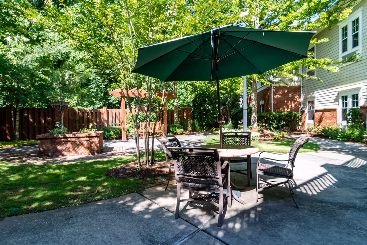 Outdoor patio area with a round table and four chairs under a large green umbrella. The patio is surrounded by greenery, including trees and bushes, with a wooden fence and a pergola in the background. Part of a building with white siding and brick accents is visible on the right side.