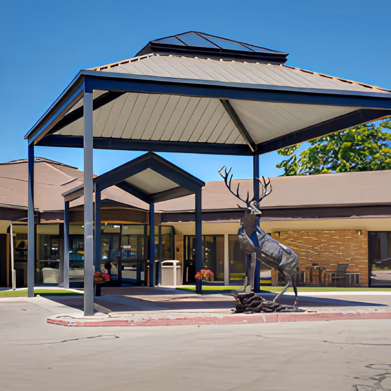 Entrance of Sandstone North Park facility featuring a covered drop-off area with a metal roof supported by steel beams. In front of the entrance is a decorative metal sculpture of a deer. The building has large glass doors and windows, with some outdoor seating visible to the right. The sky is clear and blue.