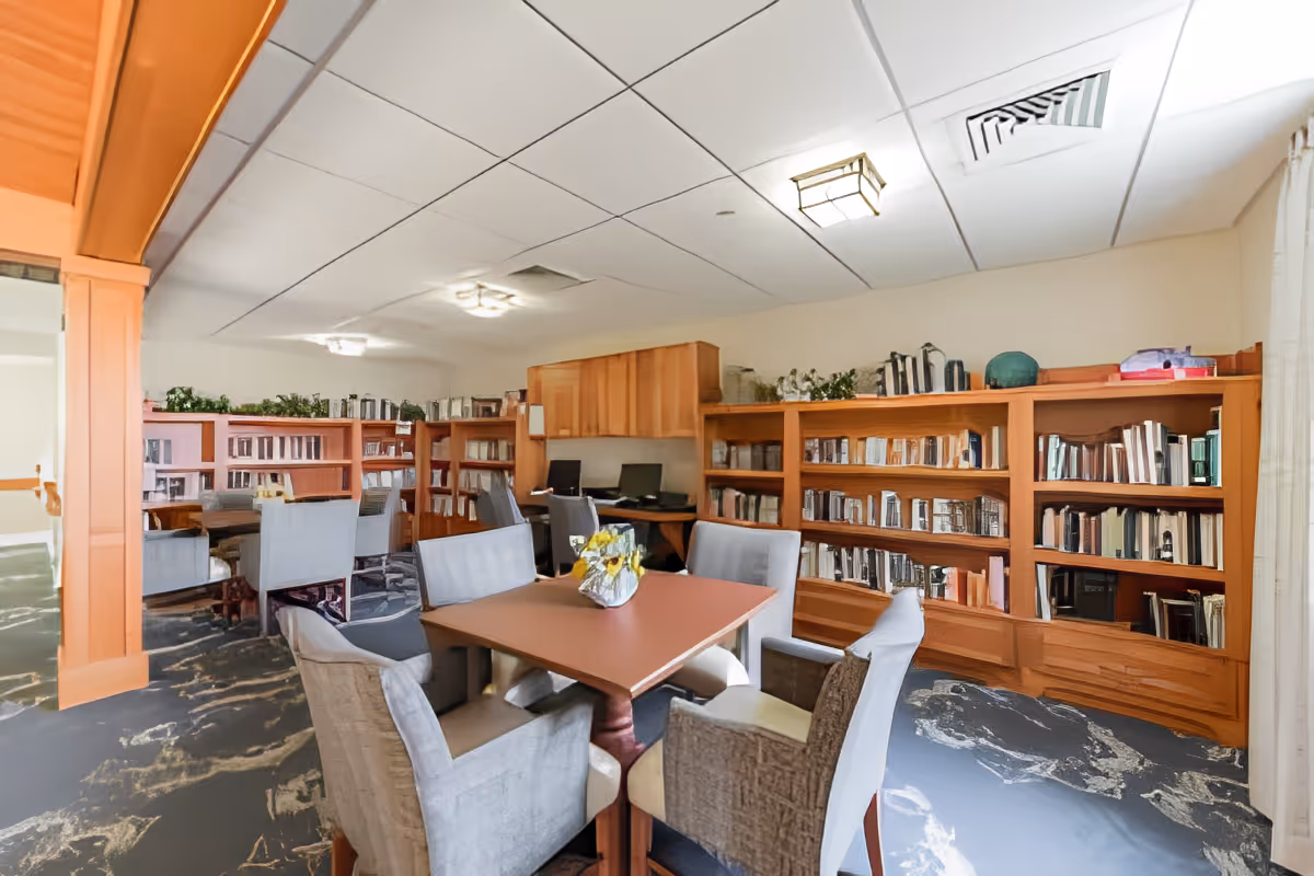 A cozy library or reading room with wooden bookshelves filled with books along the walls, a square table with a flower centerpiece, and several upholstered chairs around the table. The room has a patterned carpet, ceiling lights, and a computer workstation in the background.