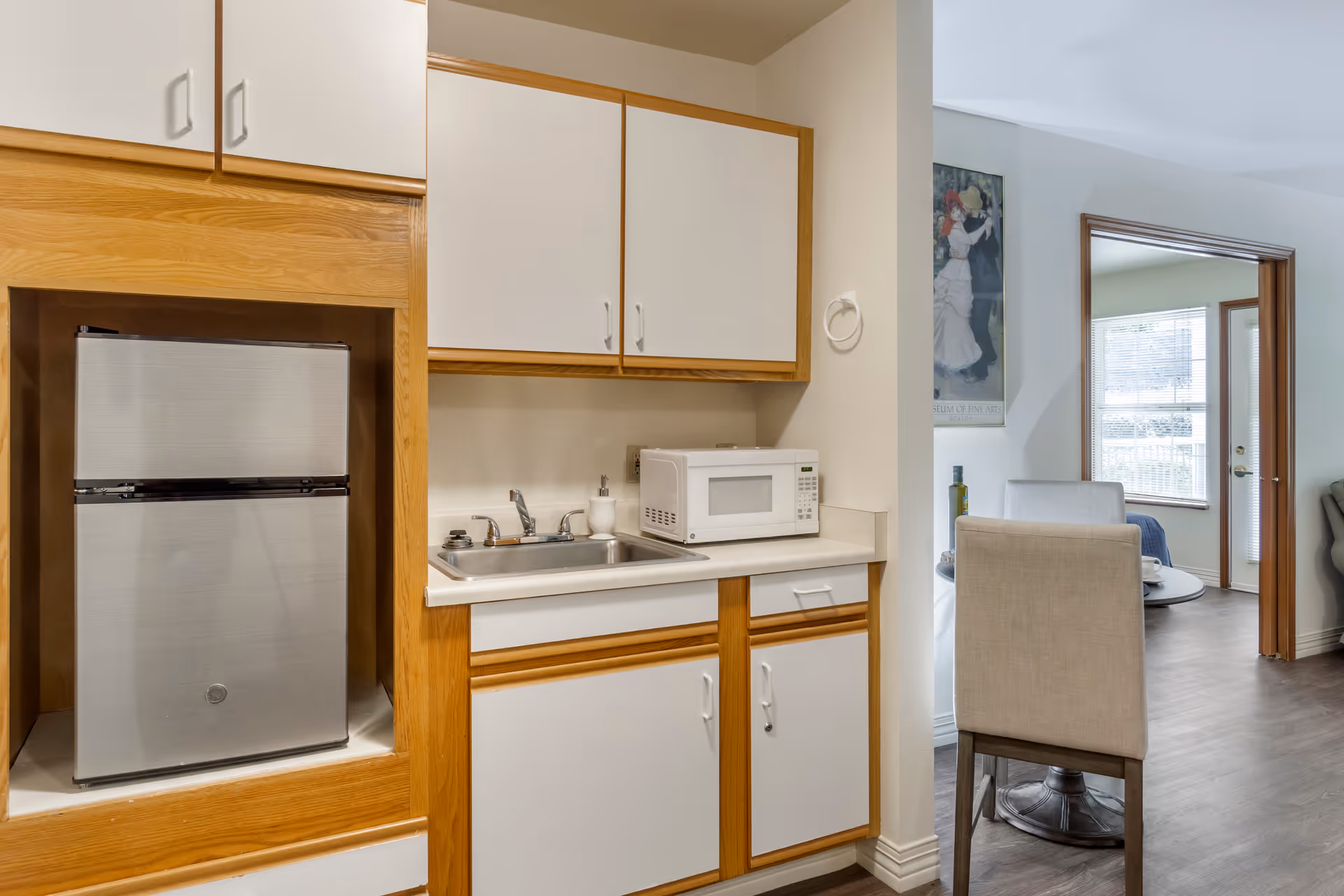 Small kitchenette featuring a mini refrigerator, sink, microwave and cabinets with a dining chair and living area visible through a doorway.