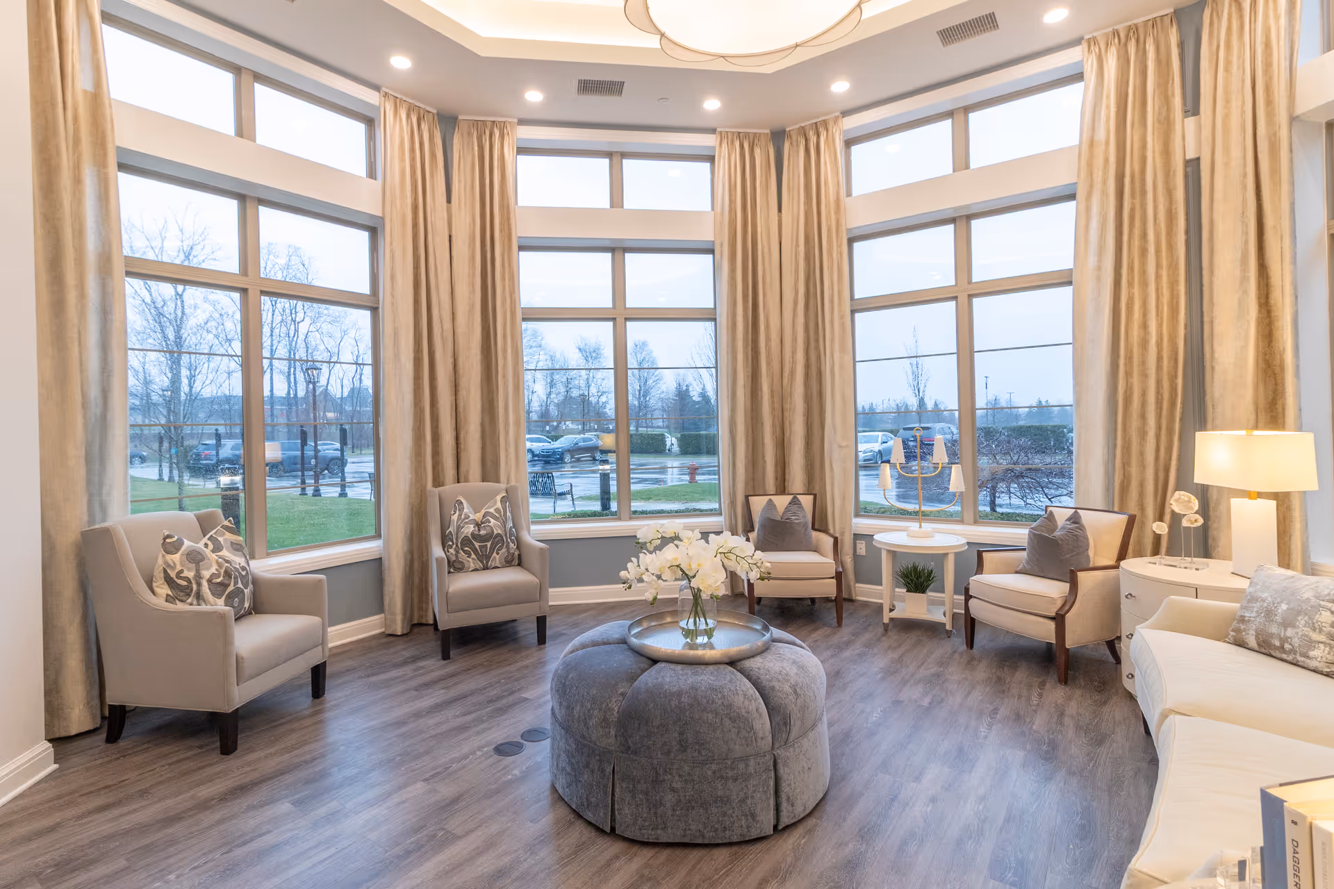 Bright communal lounge with armchairs arranged around a round upholstered ottoman and a vase of flowers in front of large bay windows with floor-to-ceiling curtains.