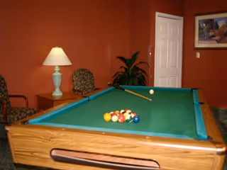 Pool table with racked billiard balls and a cue in a warm-colored recreation room with chairs, a lamp, plant, and wall art.