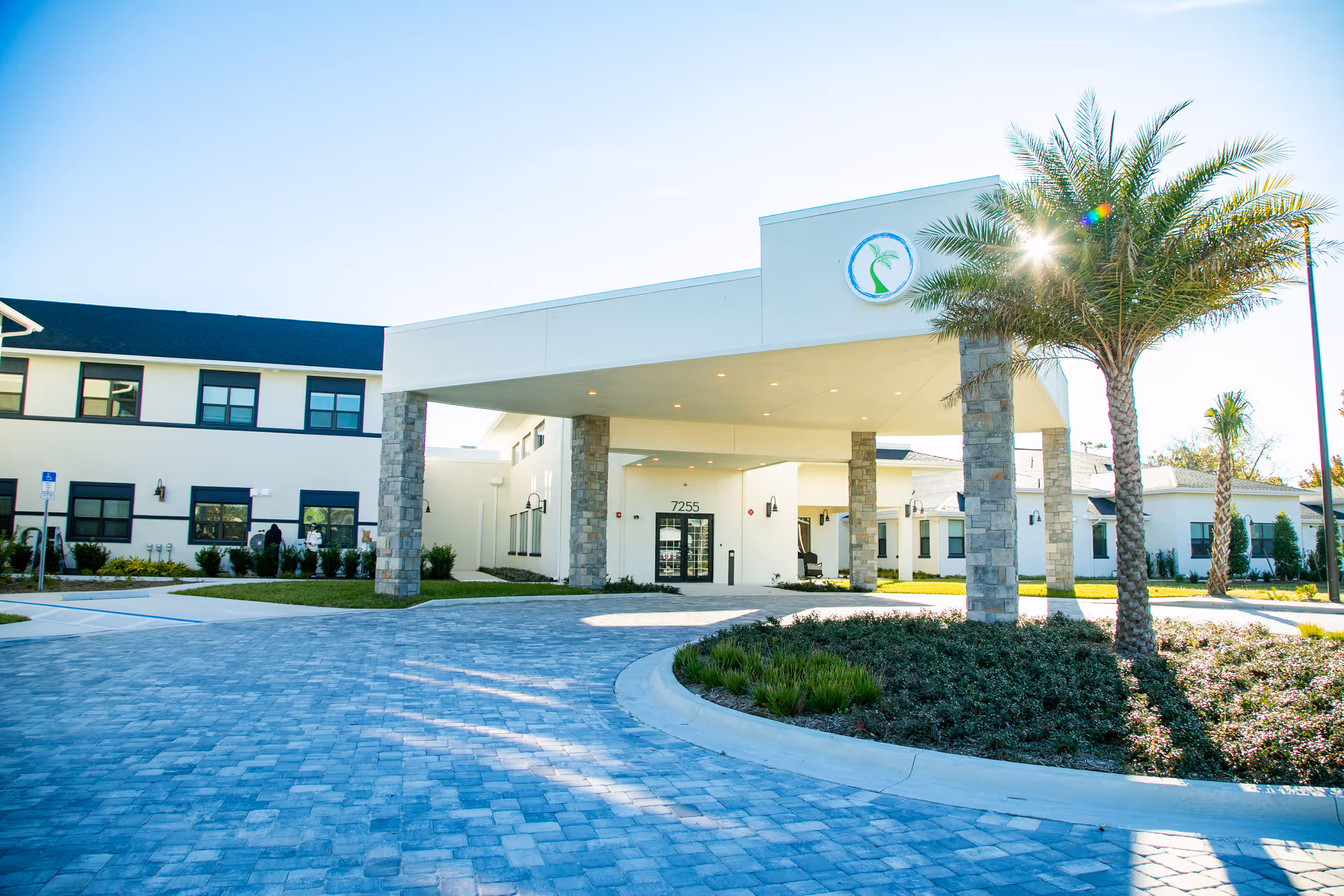 Front exterior view of Strive at Fern Park senior living community building with a covered entrance supported by stone pillars, palm trees, and a paved driveway under a clear blue sky.