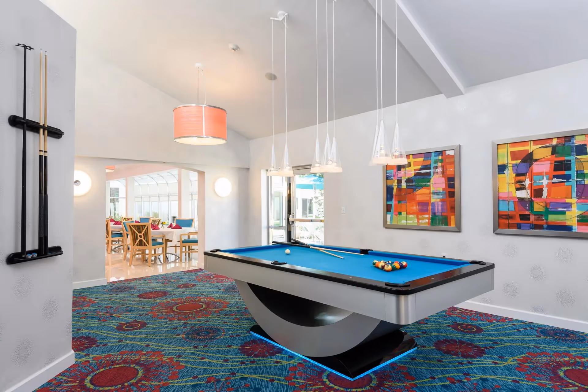 A modern game room featuring a pool table with blue felt and a set of billiard balls arranged on it. The room has colorful abstract paintings on the white walls and pendant lights hanging above the pool table. There is a vibrant patterned carpet with red and blue floral designs. In the background, there is an adjoining dining area with wooden chairs and tables, visible through an open archway.
