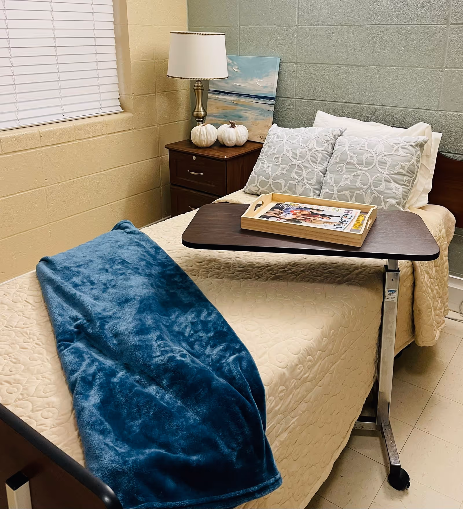 A neatly made single bed with a beige quilted bedspread, two decorative pillows, and a blue plush blanket draped across the foot of the bed. A wooden overbed table holds a tray with magazines. Next to the bed is a wooden nightstand with a brass lamp, two white decorative pumpkins, and a painting leaning against the wall. The room has beige and green painted cinder block walls and a window with closed blinds.