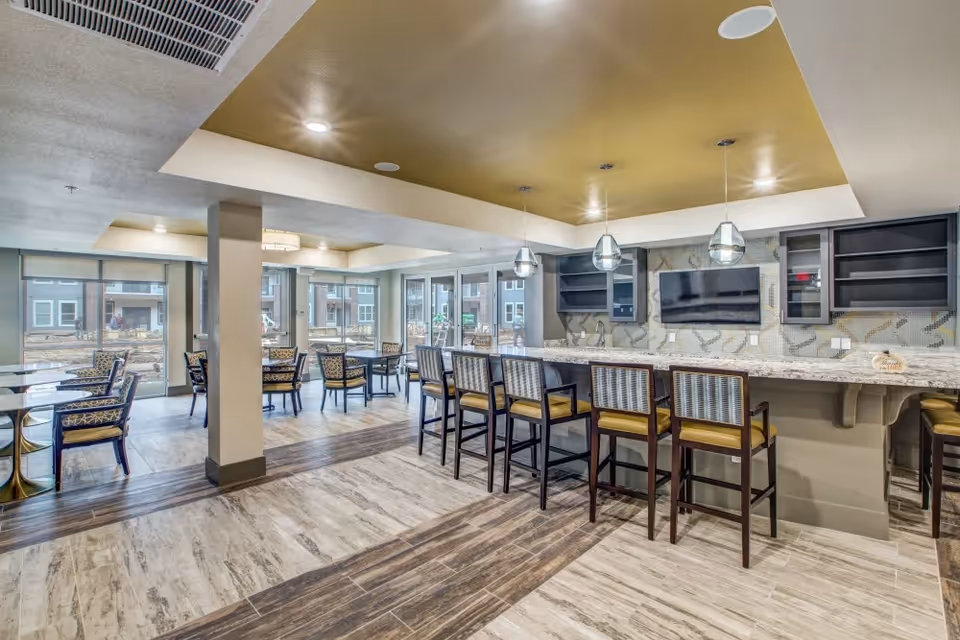 A modern dining area in a senior living facility featuring a long counter with six high chairs, pendant lights hanging above, and a wall-mounted TV. In the background, there are multiple tables and chairs near large windows that allow natural light to fill the room.
