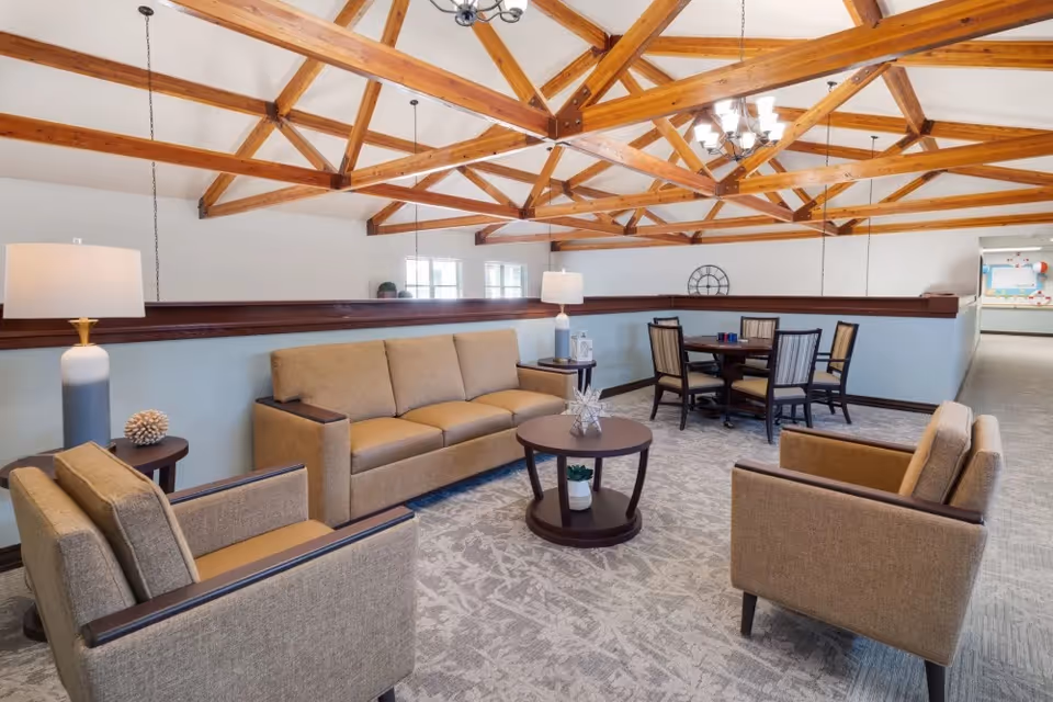 A spacious common area with beige sofas and armchairs arranged around a round wooden coffee table. Behind the seating area is a round dining table with six chairs. The room features a high ceiling with exposed wooden beams and pendant lights hanging down. The walls are painted light blue with a dark wooden trim, and the floor is carpeted with a patterned design.