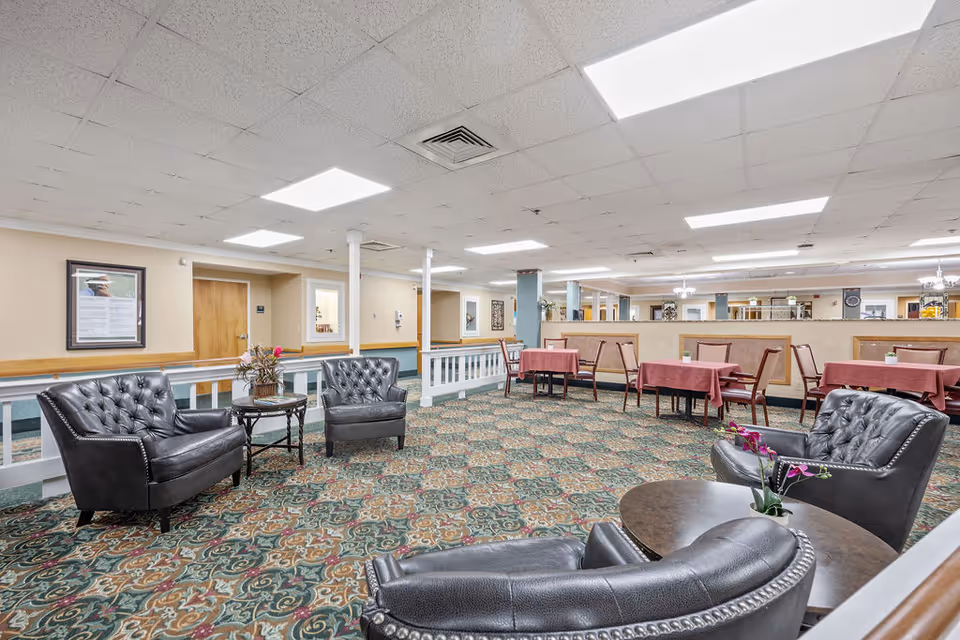 A spacious common area in a senior living facility featuring patterned carpet flooring, several black leather tufted armchairs arranged around small round tables with flower arrangements, and dining tables with pink tablecloths and wooden chairs in the background. The ceiling has recessed lighting and there are framed pictures on the beige walls.