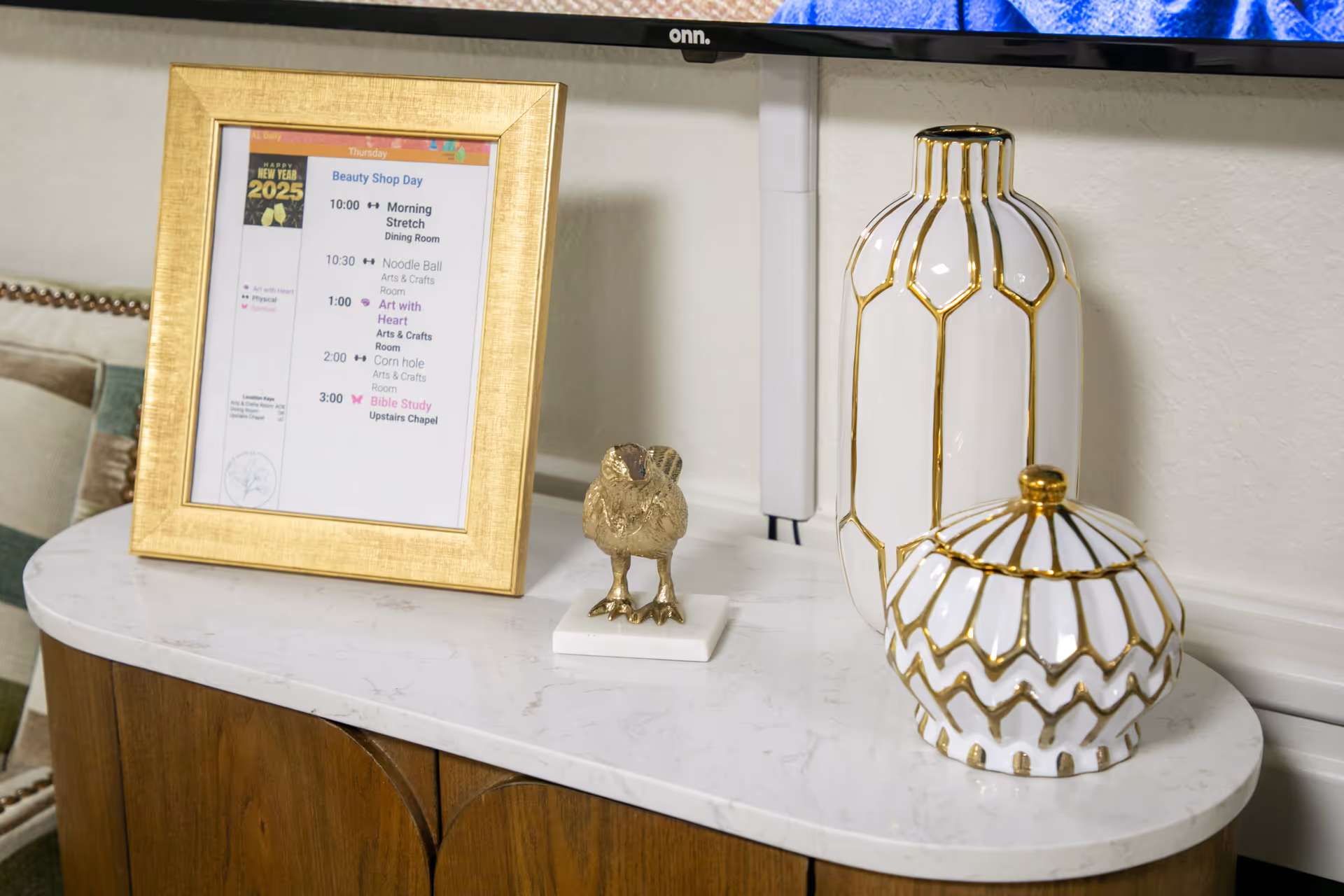 Marble-topped wooden console holding a gold-framed schedule, a small bird statue, and two white-and-gold decorative vases beneath a mounted TV.