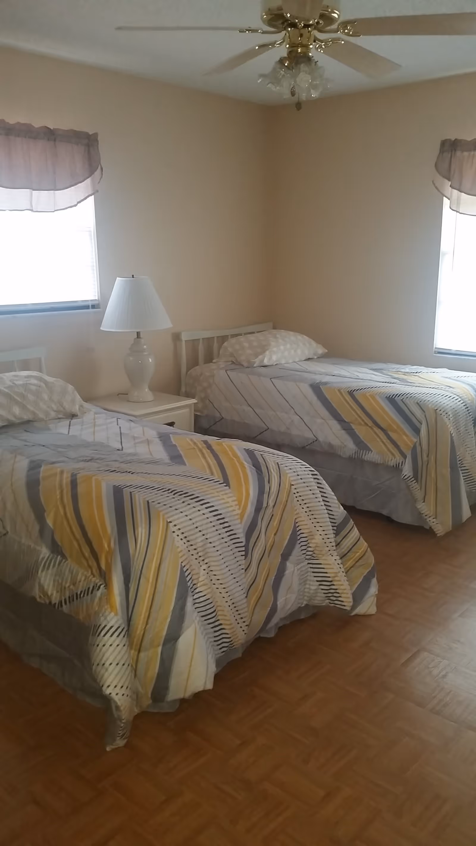 A simple bedroom with two twin beds dressed in striped bedding, a nightstand with a lamp, a ceiling fan, and two windows.