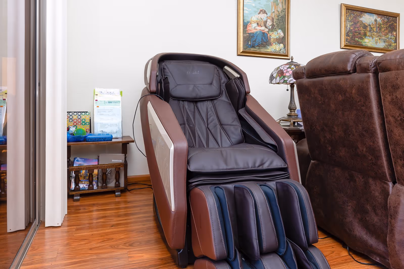 A modern massage chair in dark brown and black leather positioned on a wooden floor next to a brown leather recliner. Behind the chair is a small wooden table with various papers and items on it, and two framed paintings hang on the white wall above.