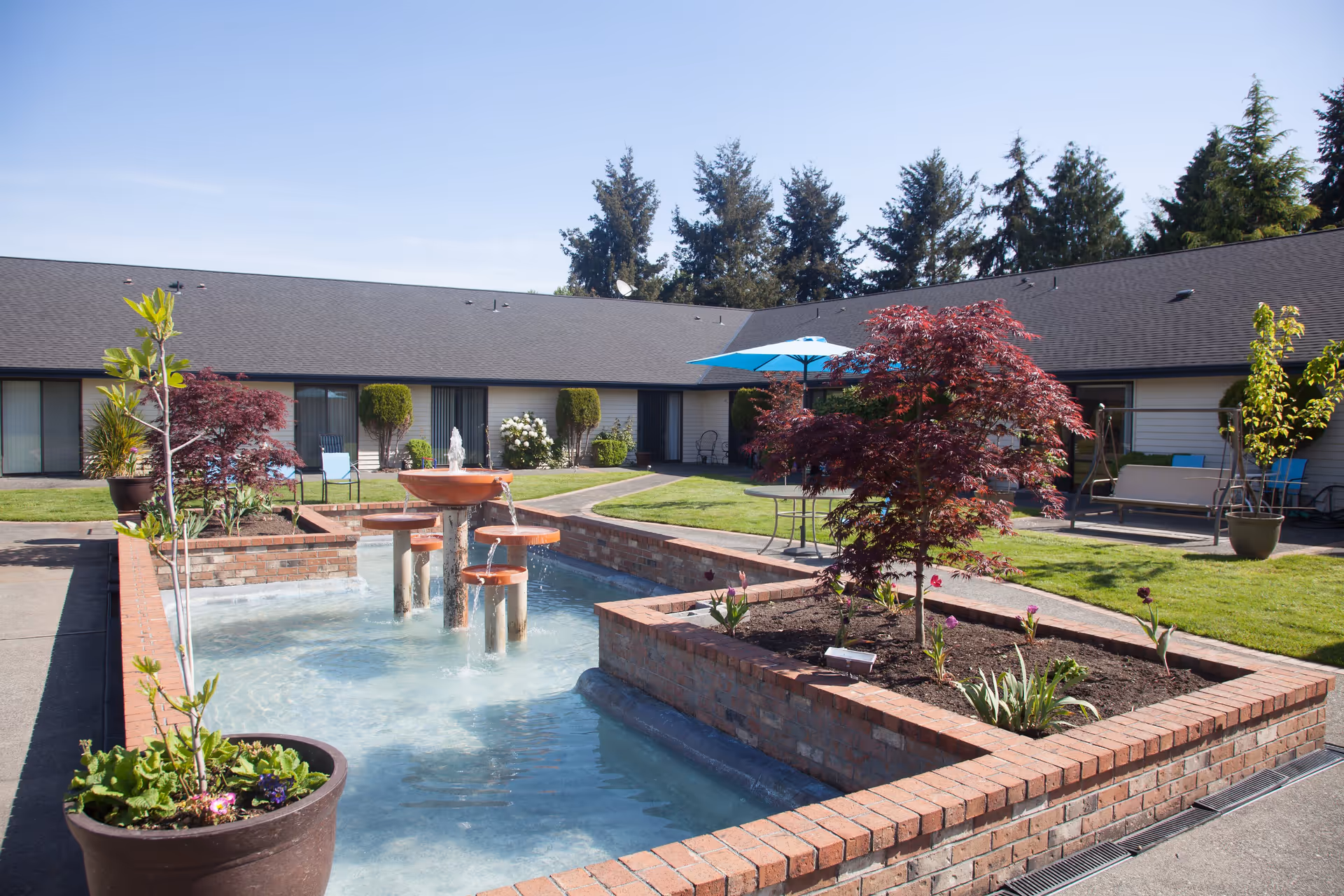 Brick fountain and landscaped courtyard with potted plants, seating and an umbrella in front of a single-story retirement community building.