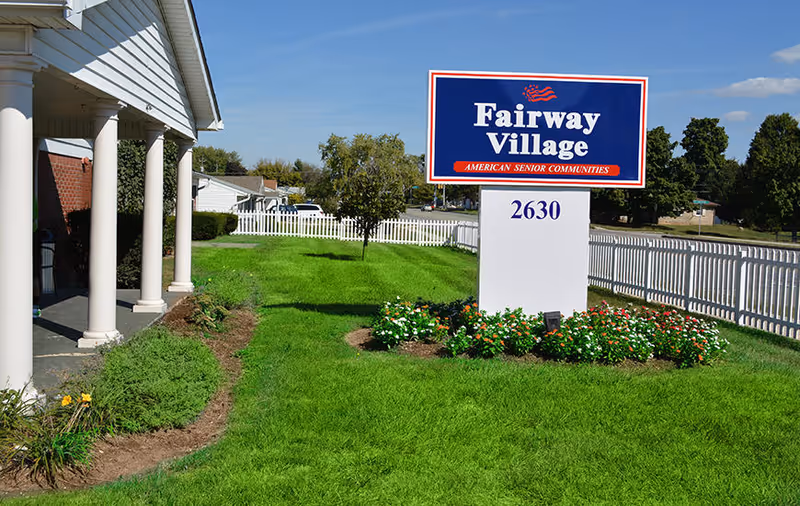 Outdoor view of Fairway Village senior living facility sign with a white picket fence, green lawn, flower beds, and part of the building with white columns under a clear blue sky.