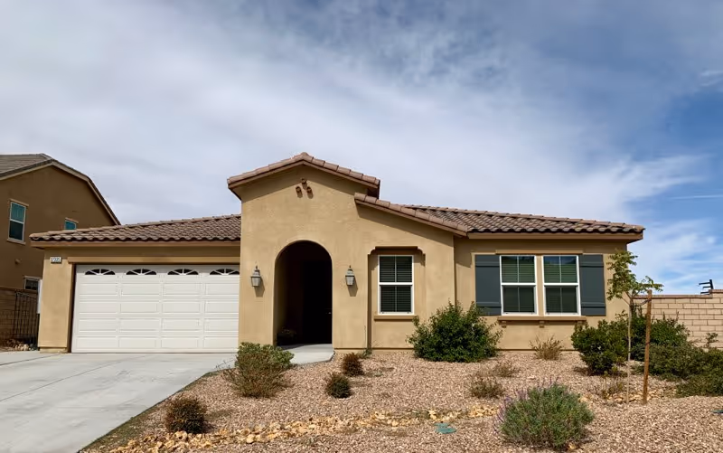 Single-story beige stucco house with a tiled roof, a two-car garage, and a small front yard with desert landscaping including rocks and shrubs under a partly cloudy sky.