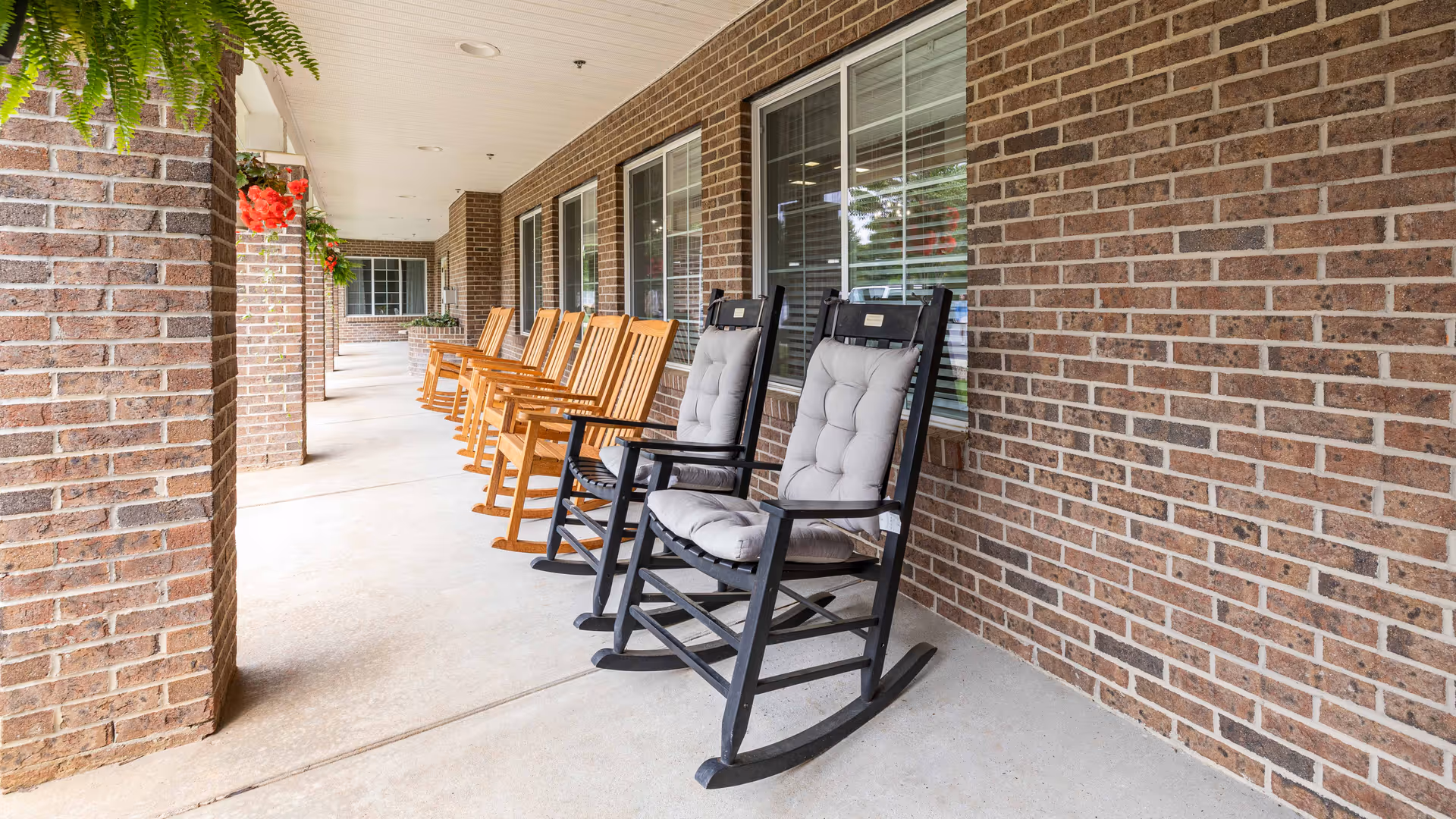 A covered outdoor patio area at Oak Park Retirement with a row of wooden rocking chairs and two black rocking chairs with gray cushions along a brick wall. Hanging plants with red flowers are visible on the left side.