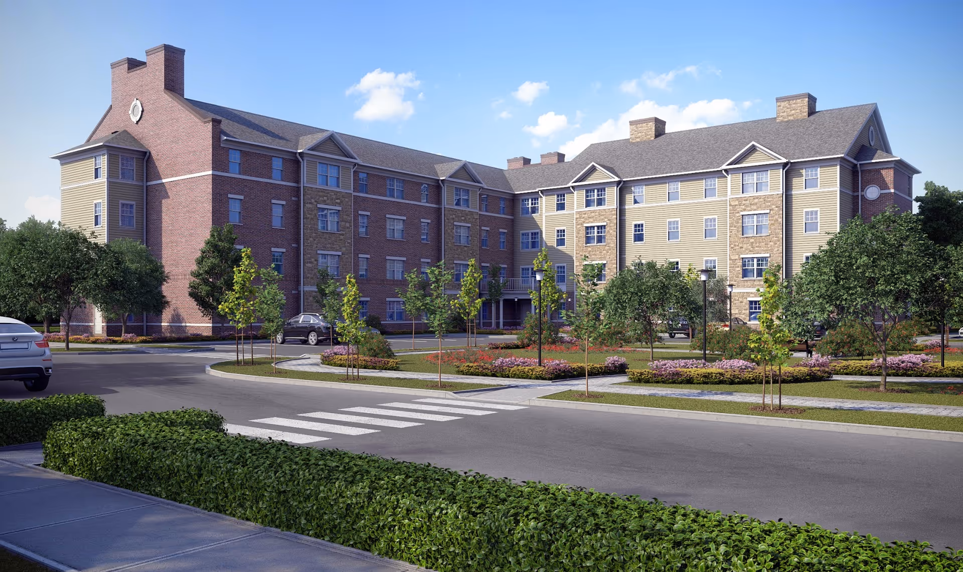 Exterior view of a multi-story senior living facility building with brick and beige siding, surrounded by landscaped greenery, small trees, and flower beds under a blue sky with some clouds. There are cars parked near the building and a crosswalk in the foreground.