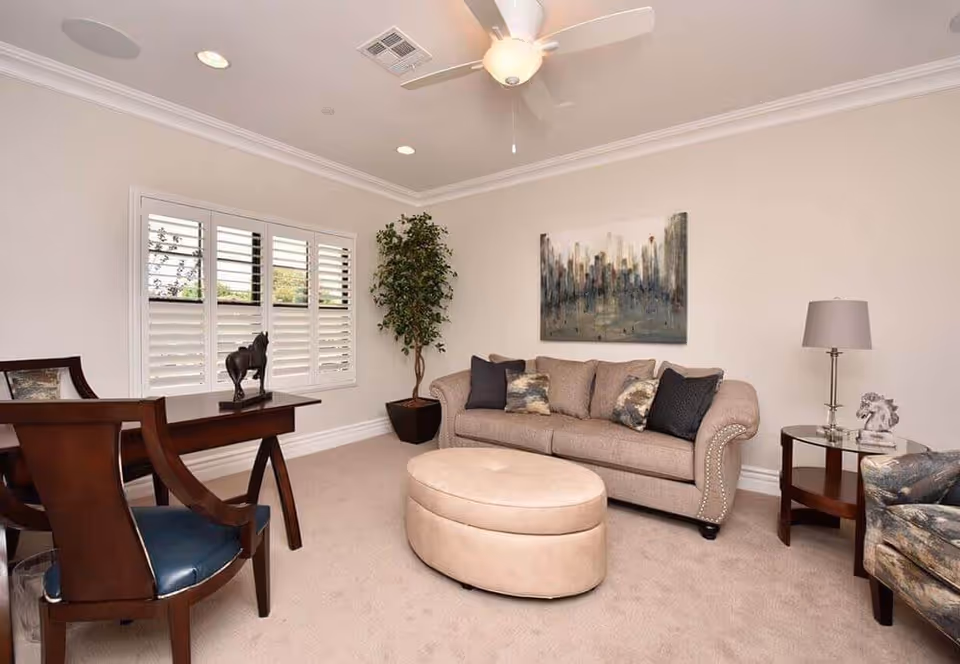 A cozy living room with a beige sofa adorned with decorative pillows, a round beige ottoman in front, a wooden desk with a chair and a horse statue, a potted plant near the window with white shutters, a side table with a lamp and a horse figurine, and a ceiling fan overhead. A cityscape painting hangs on the wall above the sofa.