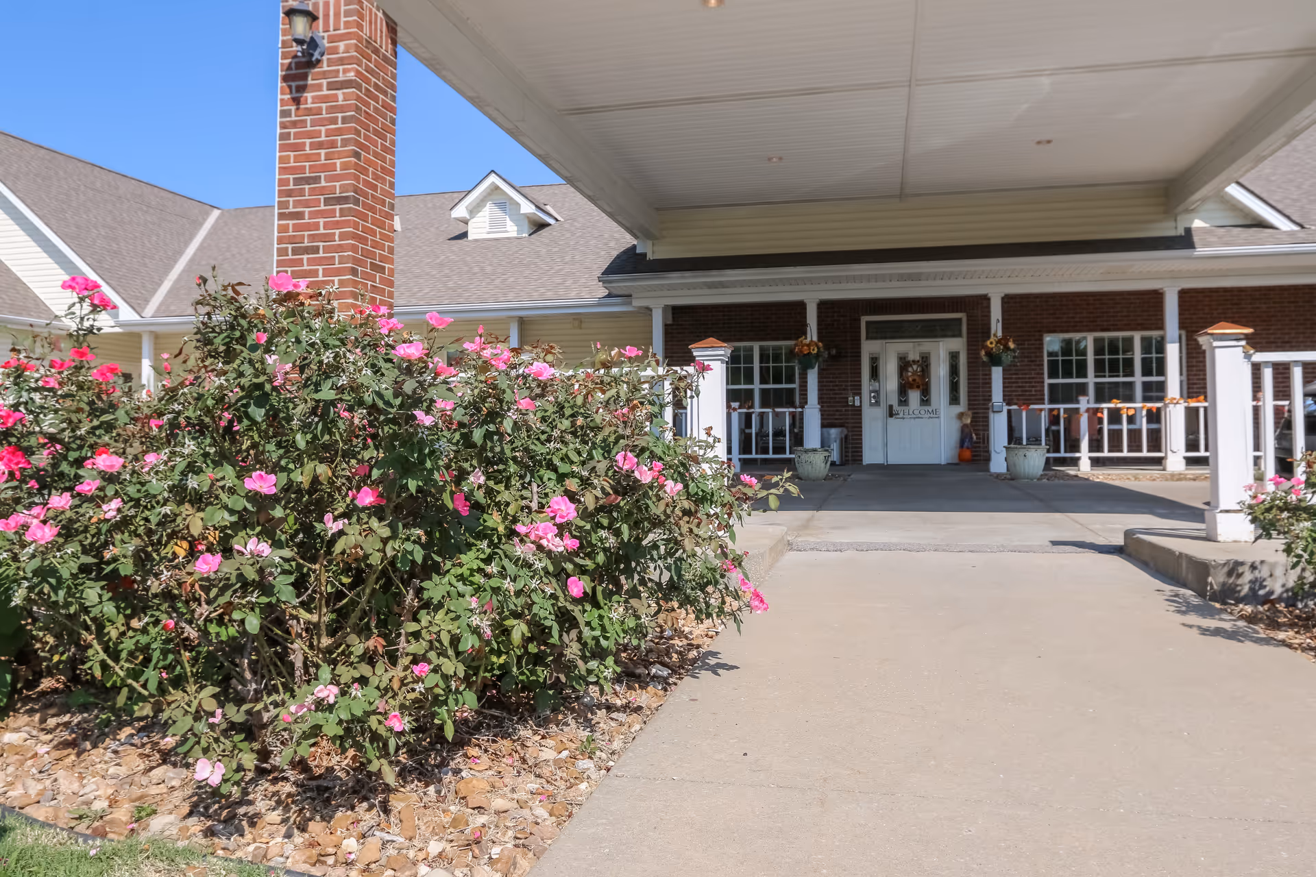 Entrance of a senior living facility with a covered driveway, brick pillars, and a garden with pink flowers in front. The building has a welcoming door with decorations and windows on either side.